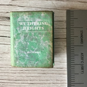 May include: Miniature book titled "Wuthering Heights" by Ellis Bell. The book has a green cover with white text and a marbled design. A ruler is next to the book, showing measurements in inches.