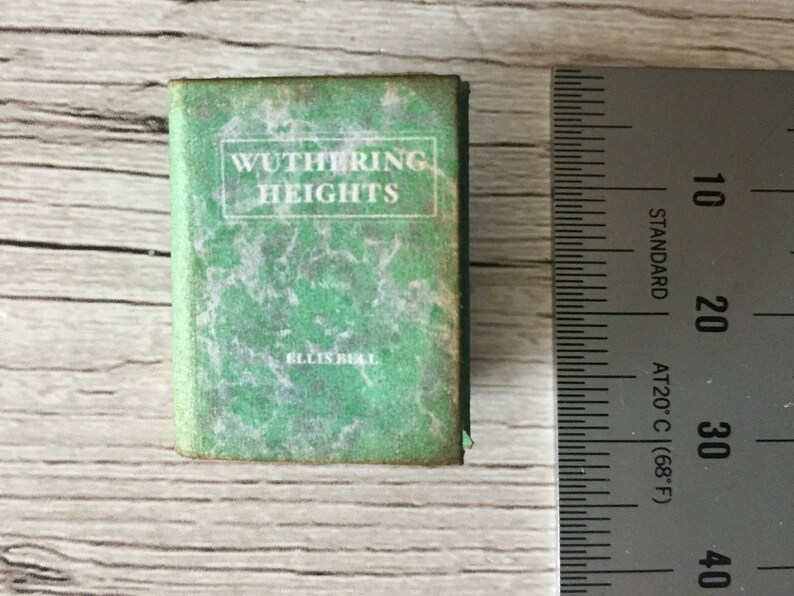 May include: Miniature book titled "Wuthering Heights" by Ellis Bell. The book has a green cover with white text and a marbled design. A ruler is next to the book, showing measurements in inches.