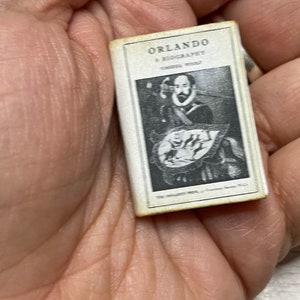 May include: Miniature book titled "Orlando: A Biography" by Virginia Woolf. The cover features a black and white portrait of a man and a decorative shield. The book is small, with a white cover and black text.