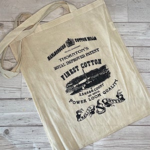 Puede incluir: Un bolso tote de algodón beige con correas largas. La bolsa presenta texto y gráficos en negro, incluyendo "Finest Cotton" y "Power Loom Quality". El diseño tiene una estética vintage.