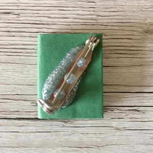 May include: A silver-toned brooch with a pin backing, featuring a glittery, light blue design with three clear, round accents. The brooch is set on a small, square, green fabric base.