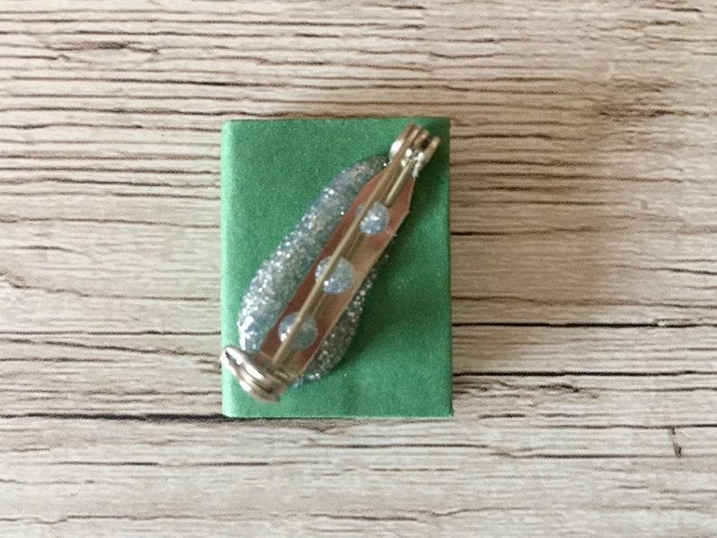 May include: A silver-toned brooch with a pin backing, featuring a glittery, light blue design with three clear, round accents. The brooch is set on a small, square, green fabric base.