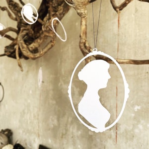 Peut inclure: Pendentif silhouette blanche d'une femme de profil, encadré dans un ovale avec un bord décoratif. Le pendentif est suspendu à un fil noir, avec d'autres ornements similaires et des branches d'arbres en arrière-plan. Thème général : décoration vintage.