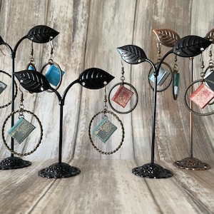 May include: Earrings displayed on decorative stands. The earrings feature miniature book charms hanging from gold-coloured hoops. The stands are black and bronze, shaped like leaves. The earrings are in various colours, including green, blue, and red.