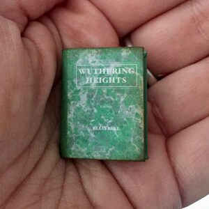 May include: A miniature book titled "Wuthering Heights" by Ellis Bell. The book has a green cover with white text and a marbled design. The small book is held in a person's hand.