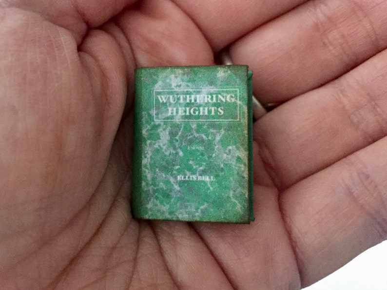 May include: A miniature book titled "Wuthering Heights" by Ellis Bell. The book has a green cover with white text and a marbled design. The small book is held in a person's hand.