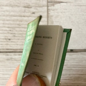 May include: A miniature book with a green cover and spine, titled "Wuthering Heights" by Ellis Bell. The book's pages are open, revealing the title and author's name in black text. The cover has a faded green color.