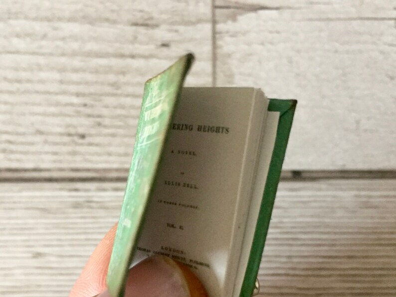 May include: A miniature book with a green cover and spine, titled "Wuthering Heights" by Ellis Bell. The book's pages are open, revealing the title and author's name in black text. The cover has a faded green color.