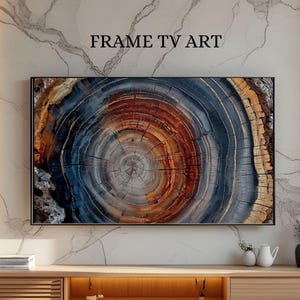 Puede incluir: Una obra de arte enmarcada para TV que presenta un primer plano de una sección transversal de un tronco de árbol. La obra de arte muestra anillos concéntricos en tonos marrones, naranjas y azules. El texto "FRAME TV ART" se muestra encima de la obra de arte. La pieza está montada en una pared de mármol.