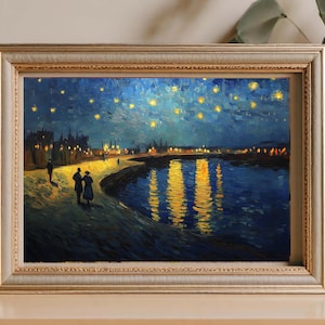 May include: A framed art print showing a night scene with a river reflecting city lights and a starry sky. The painting has a gold frame and an impressionistic style. The artwork creates a sense of peace and beauty.