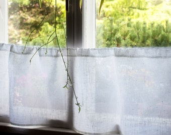 May include: White sheer curtains hang in a window with a view of green trees and foliage outside. The curtains are made of a lightweight fabric with a subtle texture.