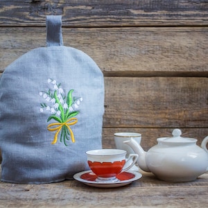 以下が含まれることがあります： 白いユリの花が刺繍されたグレーのリネン製のティーコージー。ティーコージーは、白いティーポットと2つのティーカップの隣の木製のテーブルの上に置かれています。