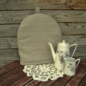 Puede incluir: Un calentador de té beige con un asa de lazo se encuentra junto a un juego de tetera y crema de porcelana blanca. La tetera y la crema tienen un diseño floral gris. Un tapete de encaje blanco se encuentra debajo del juego de té.