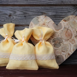 Puede incluir: Tres bolsas de lino amarillo con cordón de ajuste y ribete de encaje blanco, sentadas en una bandeja de madera en forma de corazón.