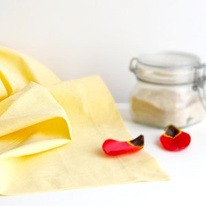 Puede incluir: Un paño de lino amarillo con dos pétalos de flores rojas sobre una superficie blanca. Un frasco de vidrio con tapa está en el fondo.