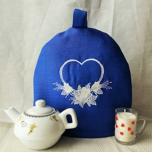 Puede incluir: Calienta-tetera de lino azul con un corazón bordado en blanco y un diseño floral. La calienta-tetera está sobre una mesa con una tetera blanca y una vela en un tarro de vidrio.
