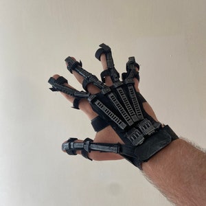 Puede incluir: Un guante impreso en 3D negro y gris con dedos articulados y una correa de muñeca.
