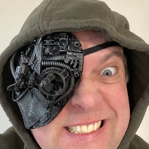 Op de afbeelding: Een persoon draagt ​​een groene hoodie met een steampunk-achtige ooglap die zijn rechteroog bedekt. De ooglap is gemaakt van metaal en heeft tandwielen en andere ingewikkelde details.