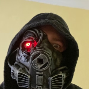 May include: A person wearing a black hooded sweatshirt with a futuristic-looking gas mask. The mask has a red light on the side and a black and silver design.