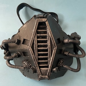May include: A black, steampunk-style gas mask with intricate details and a series of tubes and pipes. The mask has a large, rectangular, central panel with a series of vertical slats.