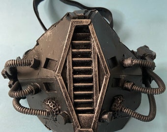 May include: A black, steampunk-style gas mask with intricate details and a series of tubes and pipes. The mask has a large, rectangular, central panel with a series of vertical slats.