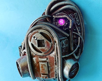 Può includere: Una maschera in stile steampunk con una finitura marrone ruggine e dettagli in metallo intricati. La maschera ha un grande occhio rotondo in metallo con una luce rosa incandescente.