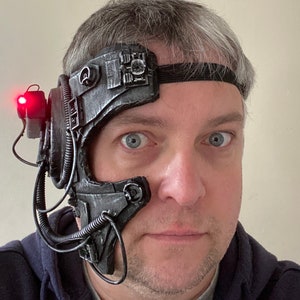 May include: A person wearing a black and silver futuristic headpiece with a red light on the side. The headpiece has a strap that goes around the head and a series of wires and tubes.