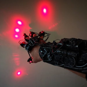May include: A black, mechanical-looking glove with red lights on the fingertips. The glove is worn on a person's hand, and the lights are pointing upwards.