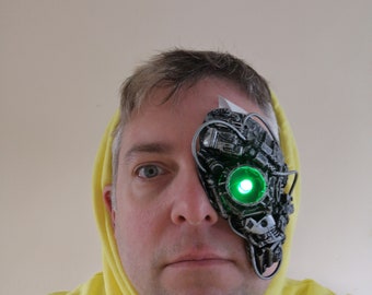 May include: A person wearing a yellow hoodie with a robotic eye prosthetic covering their right eye. The prosthetic eye has a green light.
