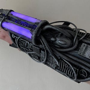 May include: A futuristic, metallic arm-mounted device with a complex design. It features two glowing purple tubes and is covered in intricate details, including wires and mechanical components. The device is held in place by a black strap.