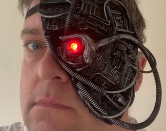 Könnte beinhalten: Eine schwarze Halbmaske im mechanischen Stil mit komplexen Schaltkreismustern, mehreren flexiblen silberfarbenen Schläuchen und einer einzelnen, markanten kreisförmigen Linse, die ein helles rotes Licht ausstrahlt, befestigt mit einem schwarzen Gummiband.