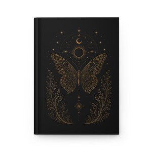 Könnte beinhalten: Ein schwarzes Notizbuch mit festem Einband und einer goldenen Linienillustration eines Schmetterlings auf der Vorderseite. Das Design zeigt himmlische Elemente wie Sonne, Mond und Sterne, flankiert von zwei symmetrischen, blättrigen Ranken.