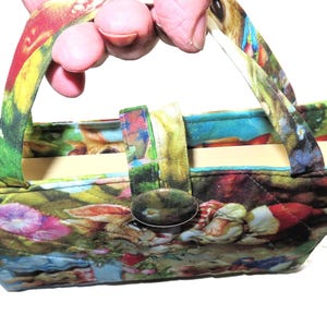 May include: A small, colorful fabric handbag with a floral and fantasy-themed print. The bag has a rectangular shape with a triangular top, a button closure, and two handles. The bag is held by a person.