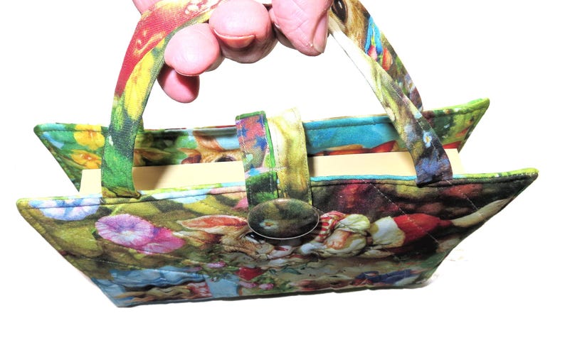 May include: A small, colorful fabric handbag with a floral and fantasy-themed print. The bag has a rectangular shape with a triangular top, a button closure, and two handles. The bag is held by a person.