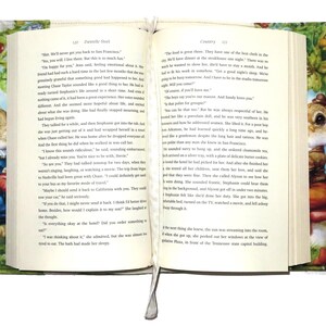 May include: Open book with a decorative cover featuring a whimsical illustration of a rabbit in a blue coat and a mushroom. The cover has a green floral pattern and integrated handles for easy carrying. A white ribbon bookmark is visible.