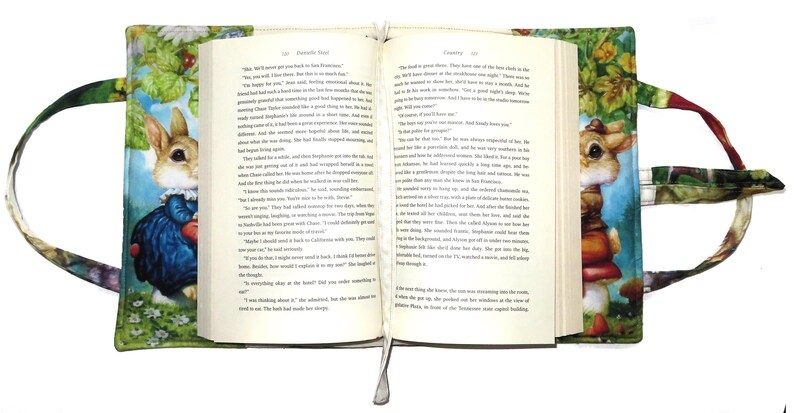 May include: Open book with a decorative cover featuring a whimsical illustration of a rabbit in a blue coat and a mushroom. The cover has a green floral pattern and integrated handles for easy carrying. A white ribbon bookmark is visible.