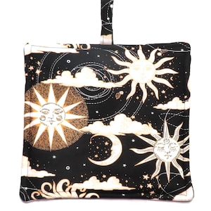 Może przedstawiać: Czarny kwadratowy przedmiot z tkaniny z niebiańskim wzorem przedstawiającym słońca, księżyce, chmury i gwiazdy w kolorze złotym i białym. Tkanina ma pętlę u góry do zawieszenia. Przedmiot jest prawdopodobnie podkładką pod garnek lub podobnym akcesorium kuchennym.