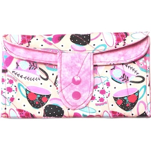 May include: A quilted wallet with a pink flap and button closure. The wallet features a cream-colored background with a pattern of pink teacups, black dots, and floral accents. The flap and button are a solid pink.