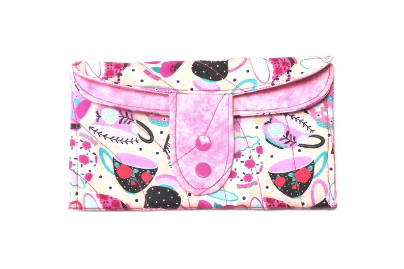 May include: A quilted wallet with a pink flap and button closure. The wallet features a cream-colored background with a pattern of pink teacups, black dots, and floral accents. The flap and button are a solid pink.