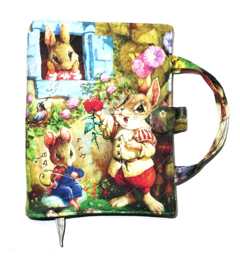 May include: Quilted journal cover featuring a whimsical scene with anthropomorphic rabbits and a mouse. The cover has a button closure and a handle. The artwork includes a rabbit holding a rose, a mouse playing a violin, and a rabbit looking out a window.