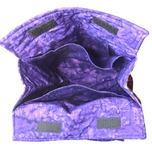 May include: A quilted fabric bag with a purple batik pattern, featuring an open top with multiple interior compartments and two black hook-and-loop fastener strips on the upper edges for closure. The interior is lined with the same purple fabric.