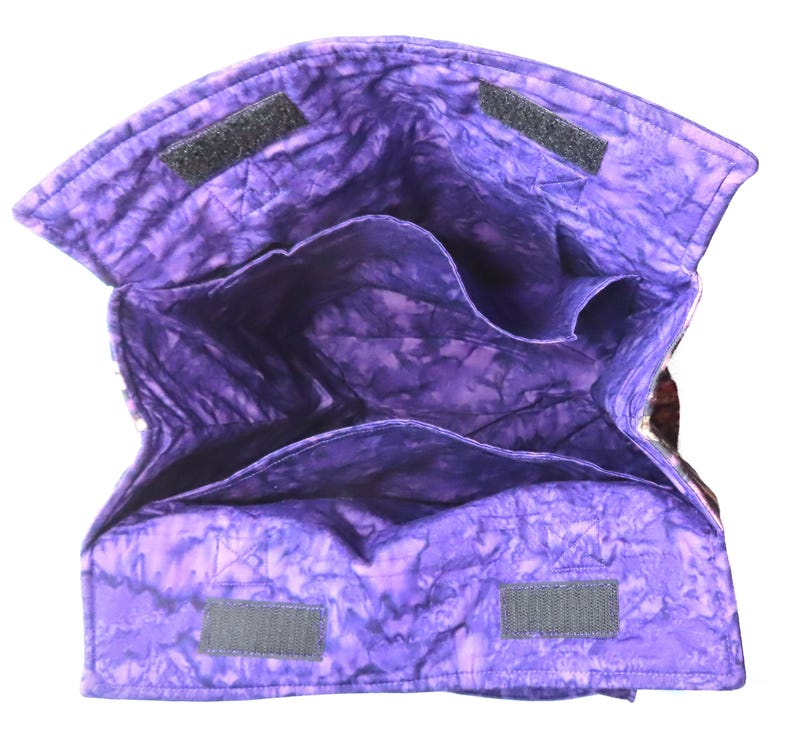 May include: A quilted fabric bag with a purple batik pattern, featuring an open top with multiple interior compartments and two black hook-and-loop fastener strips on the upper edges for closure. The interior is lined with the same purple fabric.