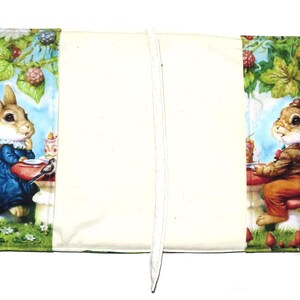May include: A fabric book cover featuring illustrations of rabbits in a garden setting. The cover has a cream-colored center with colorful panels on each side, depicting rabbits at a table with food and berries. It has handles.