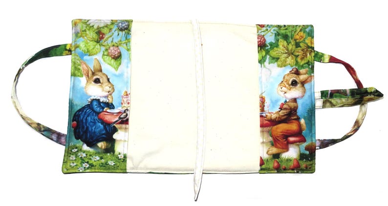 May include: A fabric book cover featuring illustrations of rabbits in a garden setting. The cover has a cream-colored center with colorful panels on each side, depicting rabbits at a table with food and berries. It has handles.