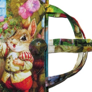 May include: A quilted fabric tote bag featuring a whimsical illustration of a rabbit in a garden scene. The bag has a button closure and a colorful handle. The rabbit is wearing a red and white outfit and holding a red rose.