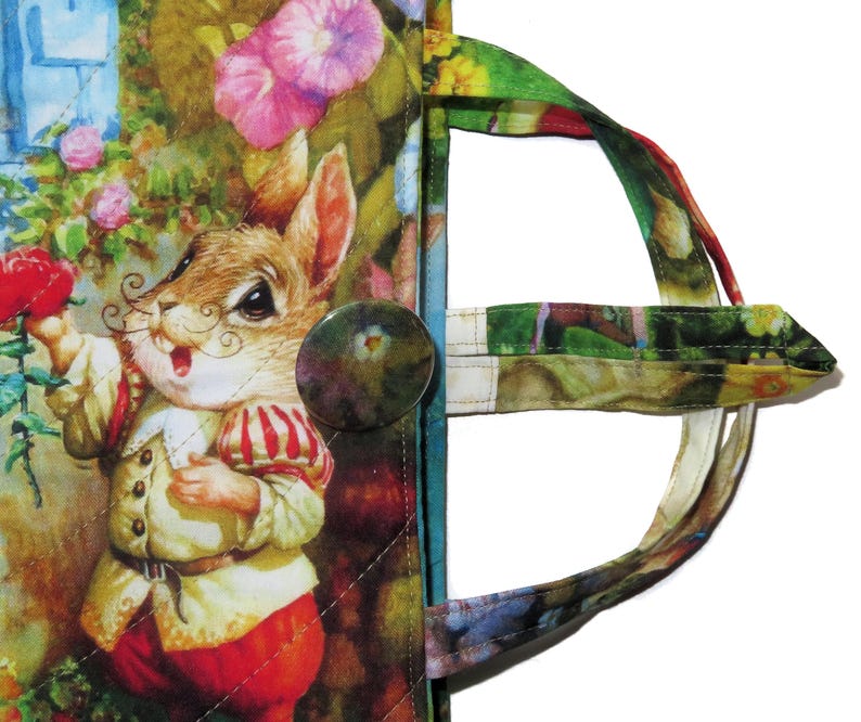 May include: A quilted fabric tote bag featuring a whimsical illustration of a rabbit in a garden scene. The bag has a button closure and a colorful handle. The rabbit is wearing a red and white outfit and holding a red rose.
