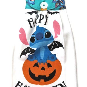 Może przedstawiać: Biały ręcznik kuchenny na Halloween z niebieską postacią z kreskówek ze skrzydłami nietoperza siedzącą na pomarańczowej dyni. Ręcznik ma nadrukowany napis "Happy Halloween". Górna klapa jest turkusowa.