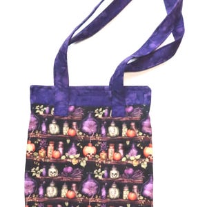May include: A fabric tote bag with two purple handles featuring a repeating pattern of shelves filled with potion bottles, skulls, and orange pumpkins against a black background. The top border of the bag is made of solid purple fabric.