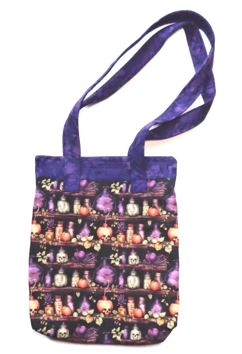 May include: A fabric tote bag with two purple handles featuring a repeating pattern of shelves filled with potion bottles, skulls, and orange pumpkins against a black background. The top border of the bag is made of solid purple fabric.