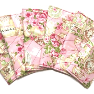May include: A set of quilted coasters with a pink floral and teacup pattern. The coasters feature a pink background with roses, teacups, and teapots. Each coaster is approximately 10 cm square.